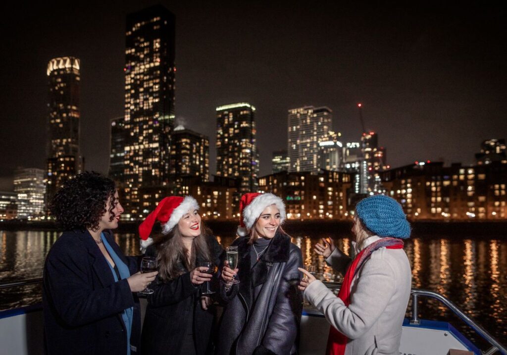 A festive Christmas Party Cruise boat sailing on the Thames at night with London landmarks illuminated, featuring a beautifully set dinner table, live entertainment, and cheerful guests