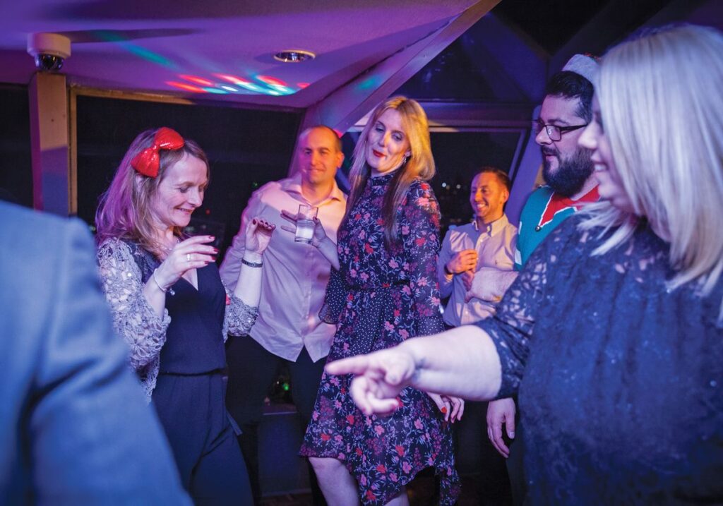 A festive Christmas Party Cruise boat sailing on the Thames at night with London landmarks illuminated, featuring a beautifully set dinner table, live entertainment, and cheerful guests